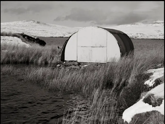 This is a black and white image. There is water, grass, tent, snow and mountains at the back.